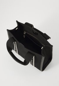 Zwarte canvas draagtas met dubbele handvatten, een interne ritszak en witte logobanden, met een gestructureerde vorm en een textuurafwerking.