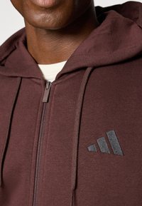 Bordeaux kleurige zip-up hoodie van zachte stof, met een gestructureerd zwart Adidas-logo op de linkerkant van de borst en een verstelbare trekkoordcapuchon.