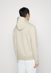 Man som bär en ljusbeige hoodie och vita byxor vänd bort mot en enkel ljus bakgrund.