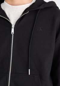 Crna zip-up trenirka izrađena od mješavine pamuka. Ima srebrni patentni zatvarač, prednje džepove i mali urezani logo na prsima.