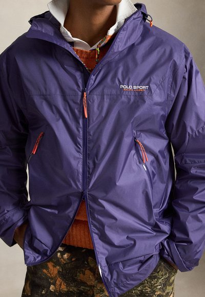 Мъж, облечен с лилаво яке Polo Sport Ralph Lauren върху оранжев пуловер и камуфлажни панталони, с ръце в джобовете.