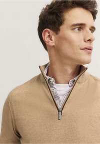 Beżowy sweter na zamek ćwierć z miękkiego materiału o prążkowanej teksturze, nałożony na białą koszulę w niebieskie paski z akcentami guzikowymi.