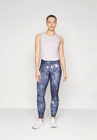 Unge kvinne står, iført en ermeløs lys grå topp, marineblå marmormønstrede tights og hvite joggesko foran en ensfarget bakgrunn.