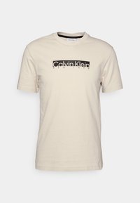 Beige tričko s krátkým rukávem a kulatým výstřihem, s černým obdélníkovým logem s nápisem "Calvin Klein New York" umístěným na hrudi.