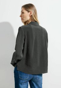 Blusa grigia a maniche lunghe con colletto, caratterizzata da una vestibilità comoda e una texture morbida, abbinata a jeans blu, vista da dietro.