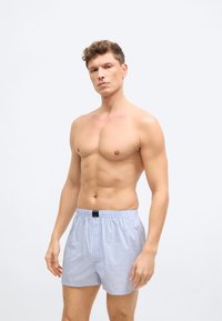 Άνδρας με κοντά σγουρά μαλλιά, φοράει ανοιχτό μπλε και άσπρα ριγέ μποξεράκια, στέκεται μπροστά σε απλό λευκό φόντο.