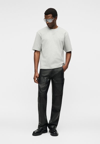 Άνδρας που φορά γκρι κοντομάνικο μπλουζάκι, μαύρο δερμάτινο παντελόνι cargo, μαύρα παπούτσια και μεγάλα ανακλαστικά γυαλιά ηλίου, στέκεται μπροστά σε απλό φόντο.