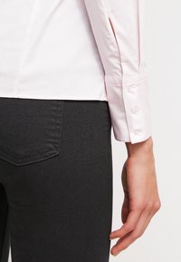 Chemise rose clair à boutons aux manches avec détail fendu, associée à un jean gris foncé. La chemise présente une texture lisse et une coupe ajustée.