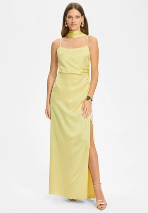 Kobieta ubrana w bladożółtą, satynową suknię bez rękawów z rozcięciem po boku, dopasowaną apaszkę na szyję, złotą bransoletkę i sandały na wysokim obcasie, stojąca na białym tle.
