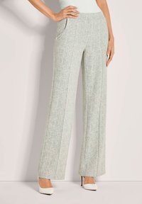 Vrouw in lichtgrijze patroon broek met hoge taille en brede pijpen, met witte puntige hoge hakken, staat met één hand op haar heup.