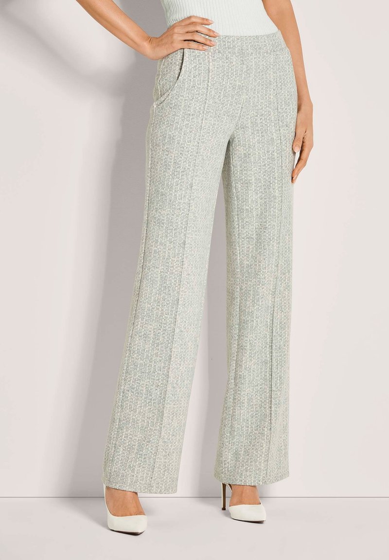 Vrouw in lichtgrijze patroon broek met hoge taille en brede pijpen, met witte puntige hoge hakken, staat met één hand op haar heup.