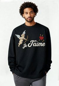 Homme aux cheveux bouclés portant un sweat-shirt noir avec un cupidon, un cœur rouge et le texte "Je T'aime", posant les mains dans les poches.