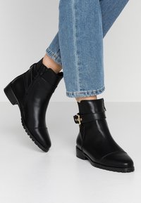 Schwarze Leder-Ankelboots mit glatter Oberfläche, Seitenreißverschluss und goldfarbenen Akzenten; niedriger, gestapelter Absatz und strukturiertes Profil.