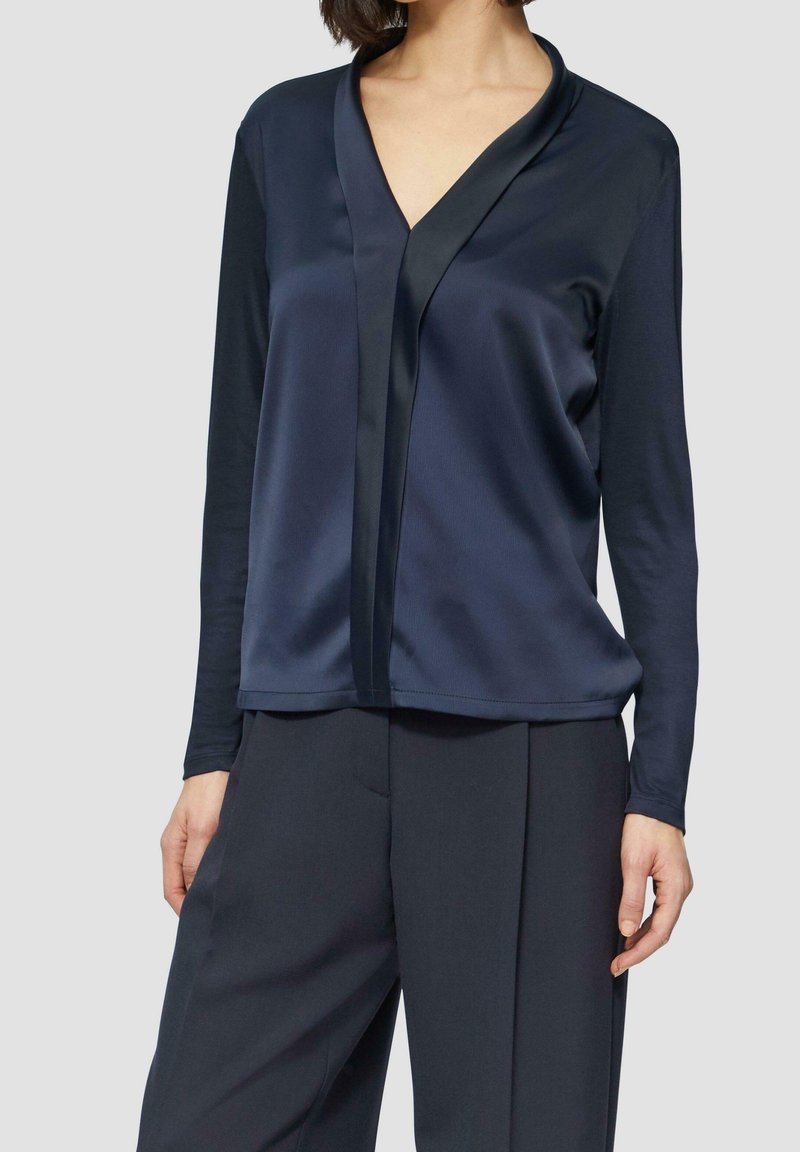 Femme portant un chemisier en satin bleu marine foncé à manches longues avec une bordure noire et un pantalon taille haute bleu marine foncé assorti sur un fond uni.
