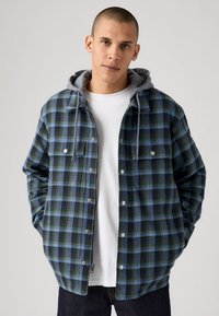 Blue and green plaid jacket with gray hoodie, button-up front, two chest pockets, and contrasting textures. White t-shirt underneath.