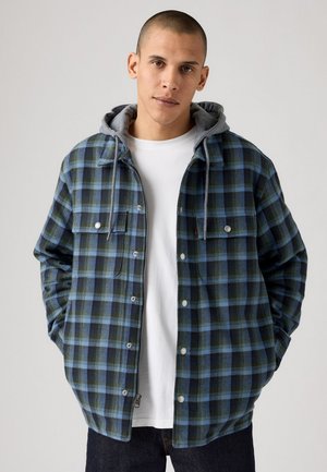 Casaco em padrão xadrez azul e verde com capuz cinza, fecho frontal com botões, dois bolsos no peito e texturas contrastantes. T-shirt branca por baixo.