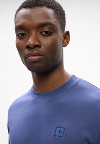 Modra mikica s krožno izrezom in rebrastimi manšetami. Ima majhen, vezen logotip v kvadratnem žepku na prsih. Gladka tekstura.