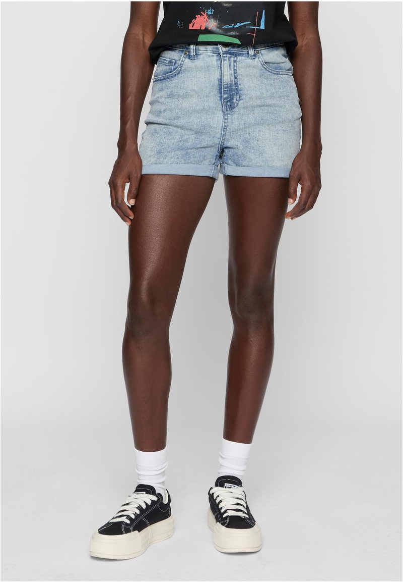 Šviesiai mėlyni džinsiniai šortai su suplėšytu kraštu, aukštu liemeniu ir sukamu kojų kraštu. Pora su juodais sportbačiais ir baltais kulkšnies kojinėmis.