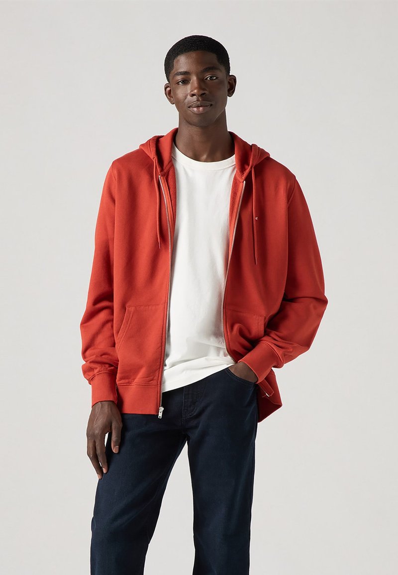 Felpa rossa con zip, dotata di lacci, tasche frontali e polsini a coste, abbinata a una t-shirt bianca e pantaloni blu scuro. Texture liscia.