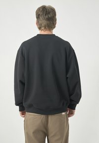 Schwarzer Sweatshirt mit entspannter Passform, Rippbündchen und runder Ausschnitt. Getragen über beigen Hosen, die ein Etikett am Bund haben.