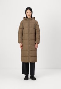 Lang, puffet brun frakk med glidelås foran, hette og elastiske mansjetter. Har et kne-langt design og er kombinert med svarte vide bukser og sko.