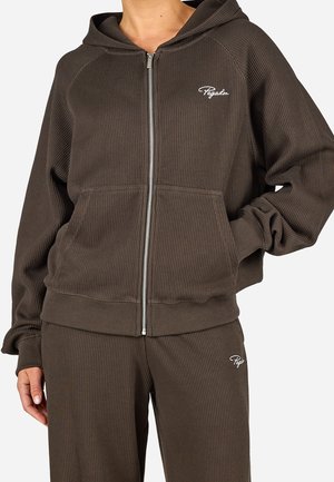 Personne portant un sweat à capuche zippé marron foncé côtelé avec poches avant et pantalon côtelé assorti avec logos blancs brodés.
