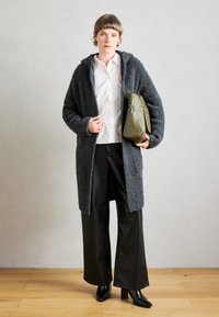 Cappotto grigio testurizzato con cappuccio, indossato sopra una camicia bianca. Abbinato a pantaloni larghi in pelle nera e stivaletti con tacco neri, mentre tiene una borsa verde oliva.