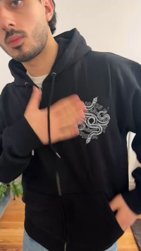 Man draagt zwarte hoodie met wit slangenontwerp, bewegingsonscherpte op hand over borst, houten vloer en planten op de achtergrond.