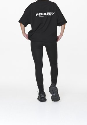 Person iført en sort oversized T-shirt med teksten "PEGADOR", sorte leggings og sorte sneakers, stående mod en hvid baggrund.