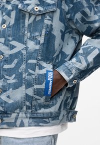Μέση ατόμου που φορεί τζιν μπουφάν με μπλε σχέδια και ετικέτα «KARL LAGERFELD JEANS» στην τσέπη, και κάτω από αυτό λευκό πουκάμισο.