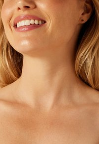 Close-up van de nek en het gezicht van een glimlachend persoon, met een gladde huid, natuurlijke tinten en studoorbellen in een minimalistisch ontwerp.