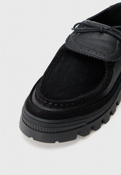 Black leather shoe featuring a textured upper, smooth accents, and a thick rubber sole with a ridged pattern. Laces and decorative stitching included.
