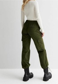 Olivově zelené cargo kalhoty s elastickým pasem a úzkými manžetami, spojené s řaseným bílým topem s dlouhým rukávem a masivními černými botami.