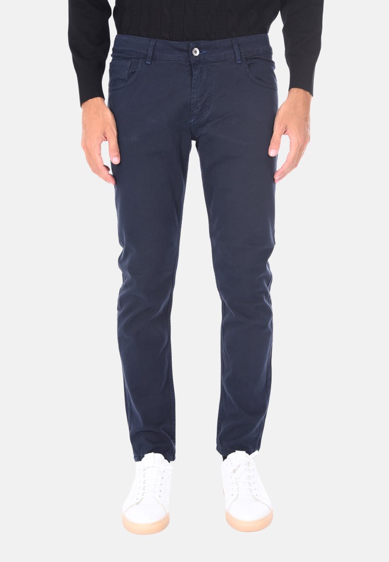 Uomo che indossa pantaloni slim-fit blu navy, una maglietta nera a maniche lunghe e sneakers bianche, in piedi con le mani rilassate ai fianchi.