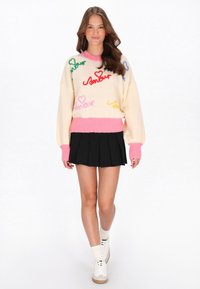 Giovane donna che cammina indossando un maglione crema con testo "amour" colorato e cuori, gonna plissettata nera, calze bianche e sneaker.