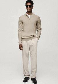 Beige ribbed sweater with a half-zip neckline, paired with light beige pleated trousers and black leather loafers. Accessories include sunglasses.