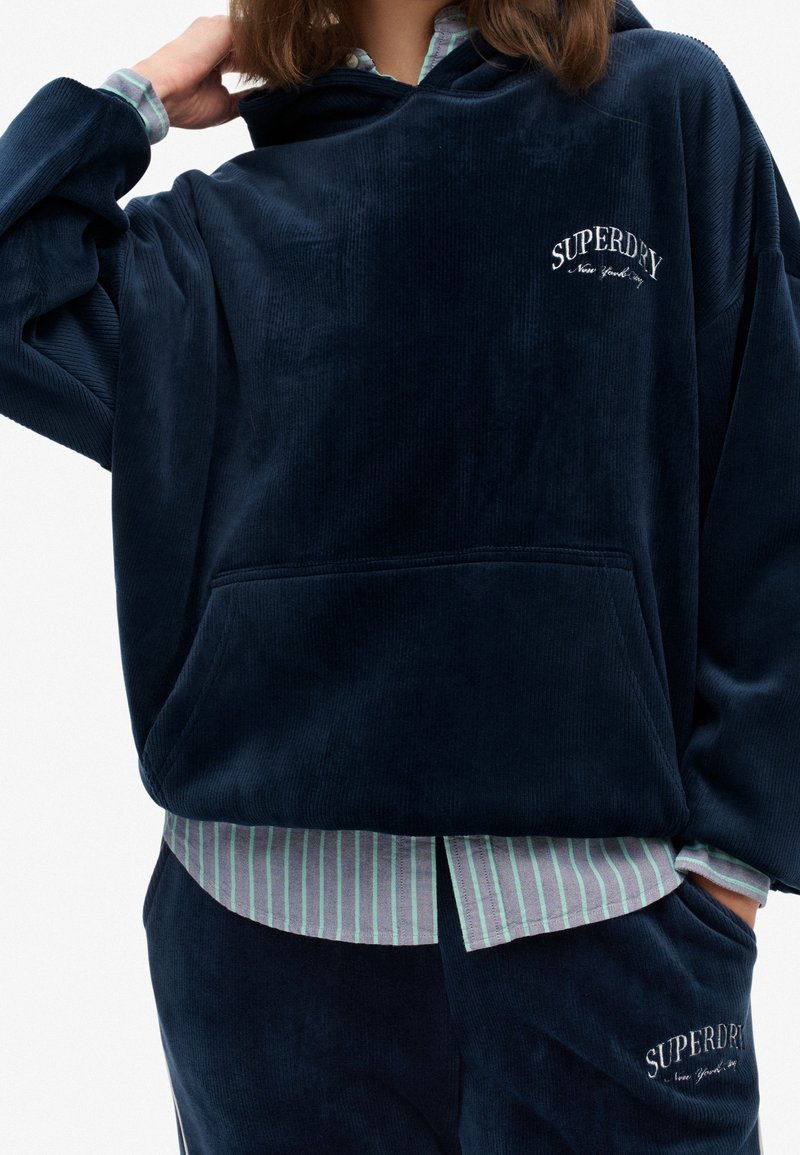 Personne portant un sweat à capuche en velours côtelé bleu marine et un pantalon assorti avec le logo blanc « Superdry », superposé à une chemise rayée boutonnée.