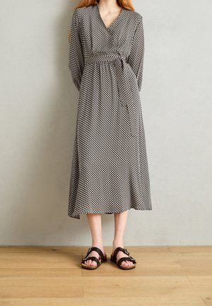 Kvinde iført en langærmet midi-kjole med sort-hvidt mønster og taljebånd samt sorte sandaler med spænde, stående på et trægulv.