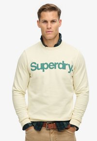 Sudadera crema con logo verde "Superdry", cuello redondo, tela texturizada. Llevada sobre una camisa de cuadros oscuros, combinada con pantalones beige y cinturón marrón.