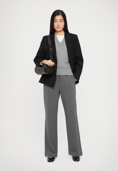 Schwarze Blazer über einem grauen Pullover, weit geschnittene graue Hose, eine runde schwarze Tasche mit einem strukturierten Tragegurt. Glatte Schuhe runden den Look ab.