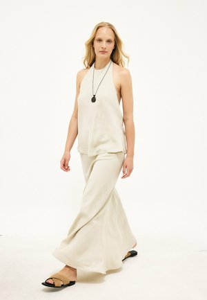 Mujer con cabello largo y rubio que lleva un top halter blanco suelto, pantalones anchos blancos, collar negro con colgante y sandalias marrones caminando.
