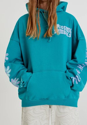 Groene hoodie met voorkantzak, roze bloemenpatronen op de mouwen en de tekst "Cosmic Symbols"; gedragen met witte broek met patroon.