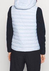 Femme portant une veste sans manches matelassée bleu clair avec capuche, par-dessus un pull noir à manches longues et un pantalon noir, vue de dos.