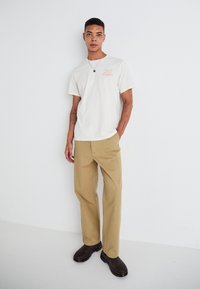 Off-white cotton t-shirt with front print, paired with beige pants and dark brown clogs. The design is relaxed, with short sleeves and a round neck.