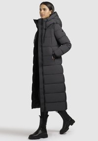 Donna che indossa un lungo cappotto in piumino nero con cappuccio e stivali neri, in piedi con una mano in tasca su uno sfondo semplice.