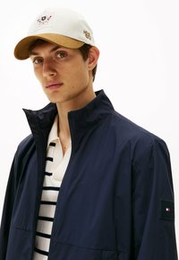 Chaqueta ligera de la marina con cuello alto, cierre de cremallera y parche con logo. Gorra blanca con visera marrón claro y el número '85' bordado. Polo a rayas debajo.