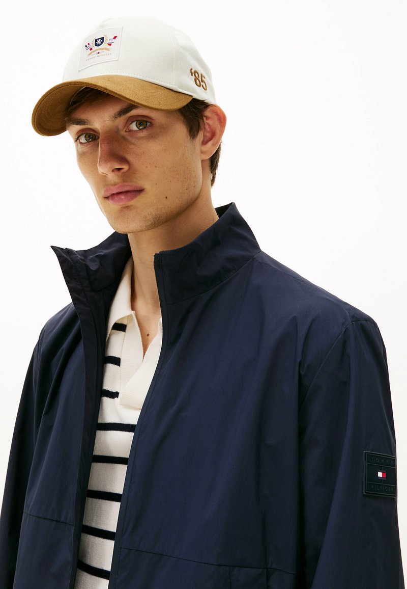 Chaqueta ligera de la marina con cuello alto, cierre de cremallera y parche con logo. Gorra blanca con visera marrón claro y el número '85' bordado. Polo a rayas debajo.