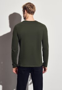 Zielona koszula z długim rękawem, wykonana z miękkiego materiału, z okrągłym dekoltem i prostym dołem. Noszona z ciemnymi spodniami, pokazana z tyłu.