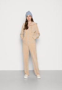 Beige samettreningdrakt med zip-opp hette og frontlommer, kombinert med matchende bukser. Modellen har på seg en lys blå strikket lue og kremfargede sneakers.
