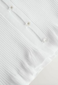 Fehér bordázott szövet, amelyen három áttetsző, virág alakú gomb van függőleges vonalban felvarrva egy fodros szegélyű ruhaélre.